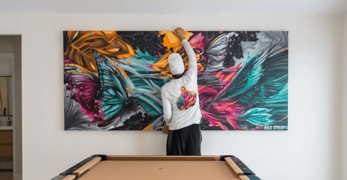 Artist Mark Francis of Bulk Styles installing a large vibrant graffiti-style canvas with butterflies and Bulk Styles branding above a pool table